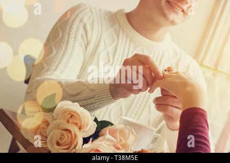 Young man putting on girlfriend finger engagement ring while having romantic dinner with flowers at cozy cafe. Engagement of young couple in love. Wed Stock Photo
