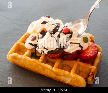 Taking a tablespoon of cream in a waffle with strawberries, whipped cream, chocolate syrup and candy colors Stock Photo