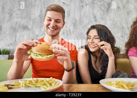 Front view of happy pair having lunch in cafe. Handsome young man and female friend talking, laughing and enjoying tasty hamburger and free potato in  Stock Photo