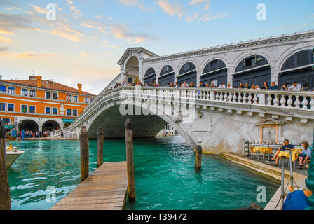 Tourists line the Rialto Bridge overlooking the Grand Canal as a sea gull perches on a pier and watches a couple dine at a cafe in Venice, Italy Stock Photo
