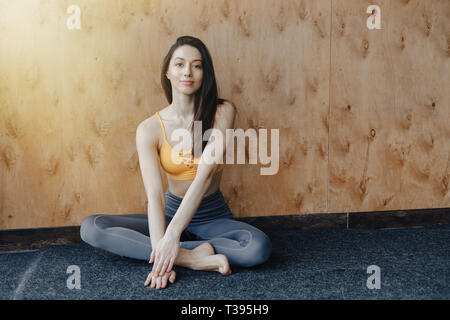 Young attractive fitness girl sitting on the floor near the window on the background of a wooden wall, resting on yoga classes at gym Stock Photo