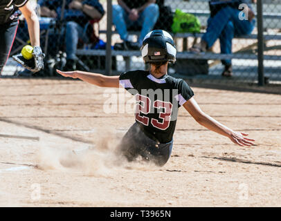 Female teenage softball player in black uniform sliding into home plate before the catcher can make the tag. Stock Photo