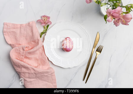 Easter table, place setting. Easter egg, pink color painted in a white plate, pink napkin, golden tableware on white marble background, vertical, copy Stock Photo