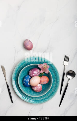 Elegance table setting with colorful Easter eggs in blue plate on white marble background, pink egg near. Concept of Holiday dinner. Flat lay, table t Stock Photo