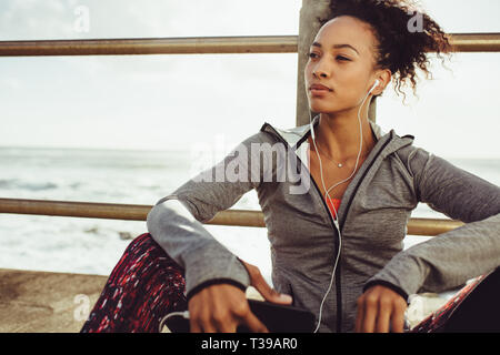 Female runner sitting outdoors and listening to music on earphones. Woman runner relaxing after her workout. Stock Photo