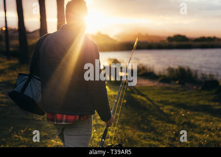 Rear view of a man walking towards a lake holding a fishing rod. Close up of a man going for fishing with sun rising in the background. Stock Photo