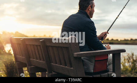 Rear view close up of a man sitting on a bench near a lake with a fishing rod. Man sitting alone near a lake and fishing. Stock Photo