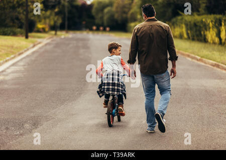 kid learning to ride a bicycle on an empty road. Rear view of a boy riding a bicycle while his father walks along with the kid.