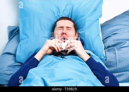 Photo of sneezing sick man in scarf and with handkerchiefs lying in bed.