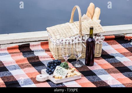 Romantic French dinner outdoors. Picnic basket, wine bottle, wine glasses, and grapes with cheese slices on checkered blanket. Red wine with grape, ye Stock Photo