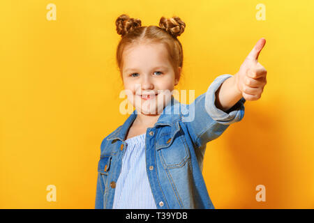 Closeup portrait of a cute attractive little child girl showing thumb up on yellow background. Stock Photo