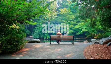 Woman sitting on a bench surrounded by green plants and tree. Stock Photo