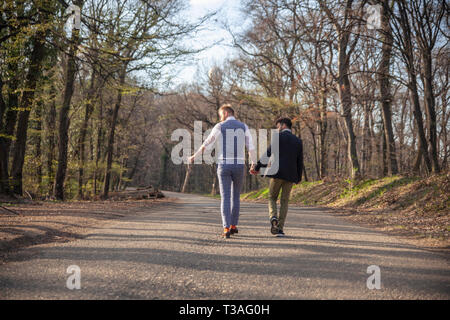 rear view, two gay man walking in forest, on asphalt road. Together holding hands. Stock Photo