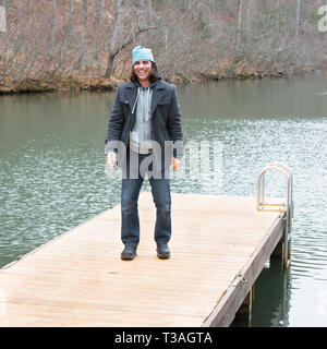 Joyous, smiling man facing the camera as he stands on a dock with lovely green lake in the background. He's wearing blue jeans, a cool jacket and cap. Stock Photo