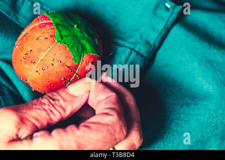 Detail of hands of senior female seamstress pinning a pin on the pincushion with green background Stock Photo