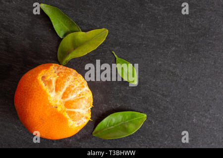 One whole fresh orange mandarine with green leaves one fruit is half peeled flatlay on grey stone Stock Photo