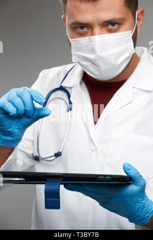 Image of doctor in mask and white coat in rubber gloves holding tablet on empty gray background Stock Photo