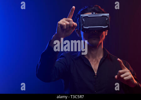 Young serious man in shirt using a virtual reality headset standing and gesturing over black background Stock Photo