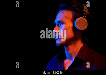 Side view of Asian mature man in shirt wearing wireless headphones and listening to music against the black background Stock Photo