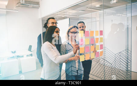 Diverse group of work colleagues smiling while brainstorming together with sticky notes on the glass wall of a modern office Stock Photo