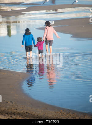 Mother and two kids walking on a beach, feet in water Stock Photo