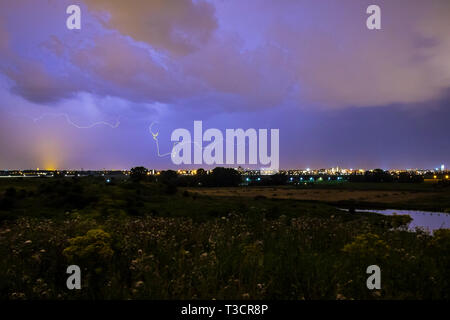 Intracloud lightning thunderbolt IC strikes at night above a city. Dramatic cloudscape thunderstorm, hills with flowers on the foreground, city on the Stock Photo