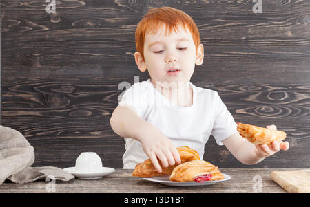 A boy while eating a dessert made from a wheat puff white bun with red cherry jam, on a black background from the boards Stock Photo