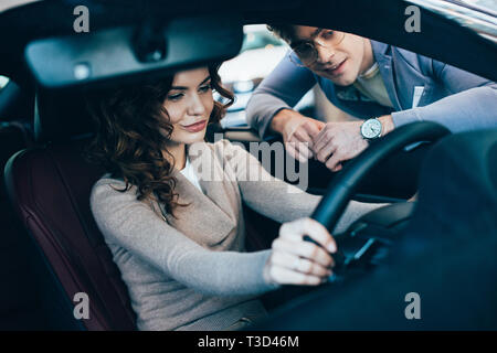 selective focus of beautiful curly woman sitting in car and holding steering wheel near cheerful man in glasses Stock Photo