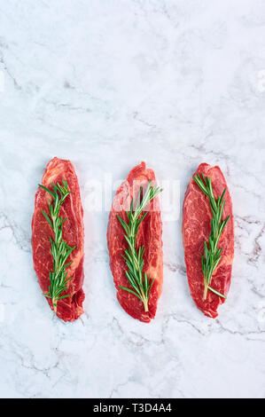 raw marbled beef steaks with green rosemary branch at white marble background. copy space Stock Photo