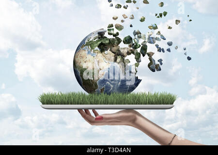 Side view of woman's hand holding tablet with green grass on screen and Earth globe on top which is dissolving into pieces that float away into sky. Stock Photo