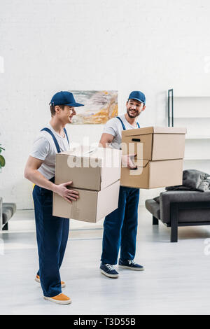 two smiling movers in uniform carrying cardboard boxes in apartment Stock Photo