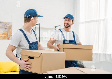 two smiling movers looking at each other while transporting cardboard boxes in apartment Stock Photo