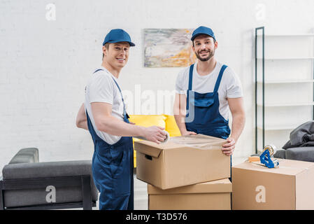 handsome smiling movers looking at camera while wrapping cardboard box with stretch film in apartment Stock Photo
