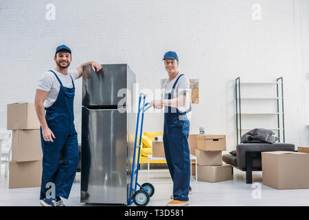 two smiling movers using hand truck while transporting refrigerator in apartment Stock Photo