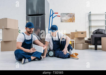 two movers using digital tablet while sitting near hand truck and refrigerator in apartment Stock Photo