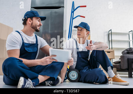 two movers in uniform using digital tablet while sitting near hand truck and refrigerator in apartment Stock Photo