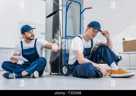 two movers using digital tablet while sitting near hand truck and refrigerator in apartment Stock Photo