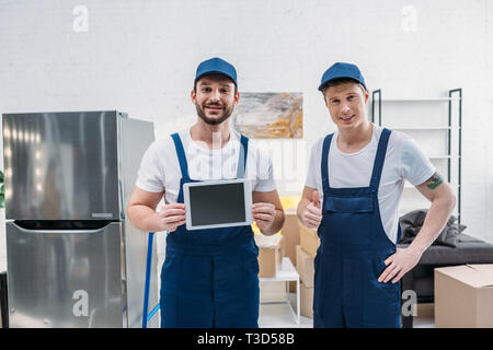 two movers presenting digital tablet with blank screen, showing thumb up sign and looking at camera in apartment Stock Photo