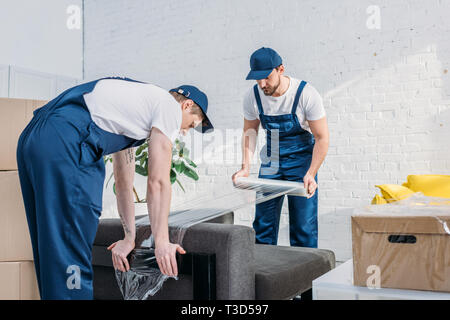 movers wrapping couch with roll of stretch film in apartment Stock Photo