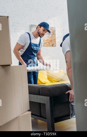 selective focus of movers wrapping couch with roll of stretch film in apartment Stock Photo