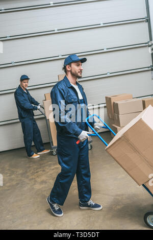 two movers in uniform transporting cardboard boxes with hand trucks in warehouse Stock Photo