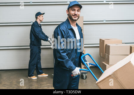 two movers in uniform transporting cardboard boxes with hand trucks in warehouse Stock Photo