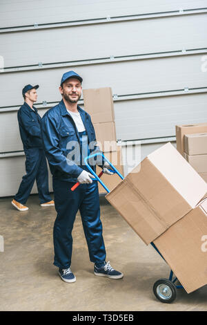 two movers in uniform transporting cardboard boxes with hand truck in warehouse Stock Photo