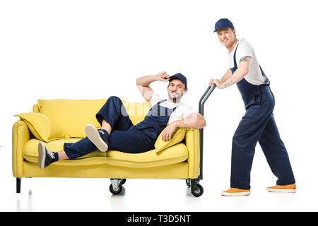 mover in uniform sitting on sofa and looking at camera while colleague pushing hand truck on white Stock Photo