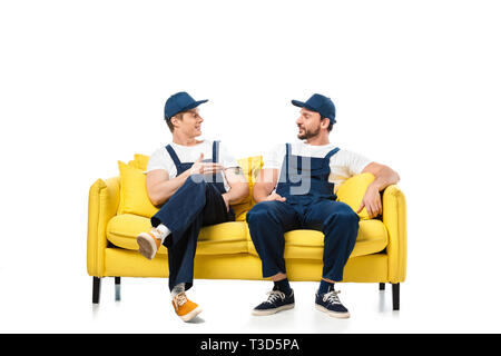 two movers in uniform sitting on yellow sofa and looking at each other on white Stock Photo