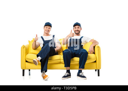 two movers in uniform sitting on yellow sofa, showing thumb up signs and looking at camera isolated on white Stock Photo