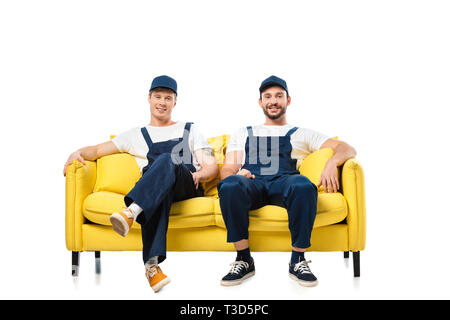 two smiling movers in uniform sitting on yellow sofa and looking at camera isolated on white Stock Photo