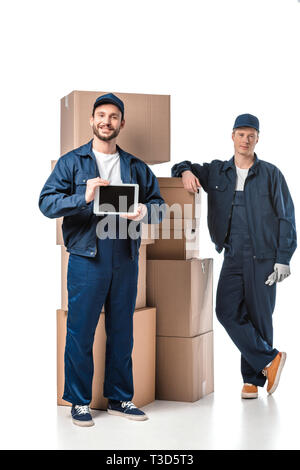 two movers with cardboard boxes looking at camera and presenting digital tablet with blank screen on white Stock Photo