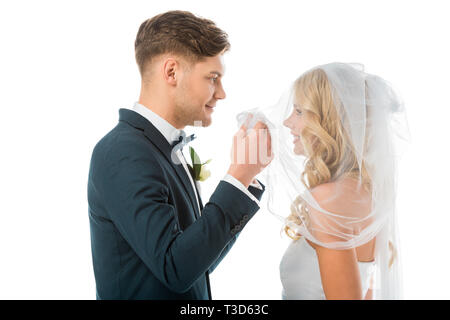 happy groom looking at smiling brides face while lifting bridal veil isolated on white Stock Photo