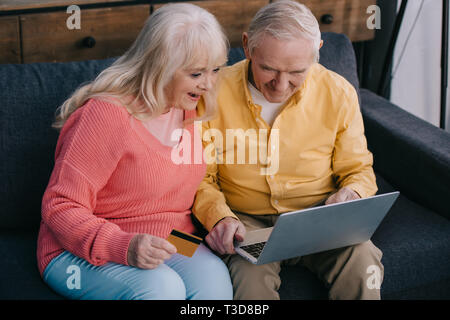 excited senior couple sitting on couch with laptop and credit card while doing online shopping at home Stock Photo